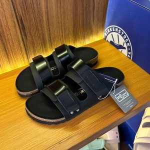 A pair of black leather sandals with a cork sole on a wooden shelf, with a blue box in the background