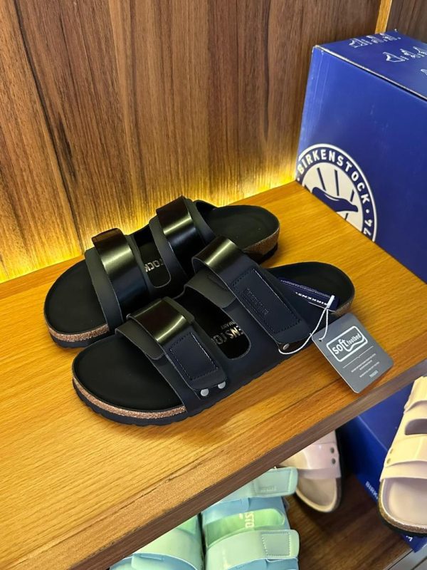 root A pair of black leather sandals with a cork sole on a wooden shelf, with a blue box in the background