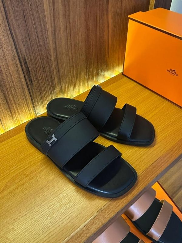 Pair of black Hermes slides on a wooden shelf, showcasing the brand's signature style and attention to detail.