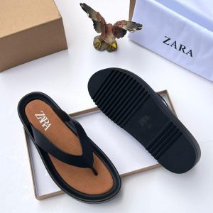 A pair of black T-strap flip flops with brown insole and rubber sole, placed on a white surface with a shoe box and bag in the background.