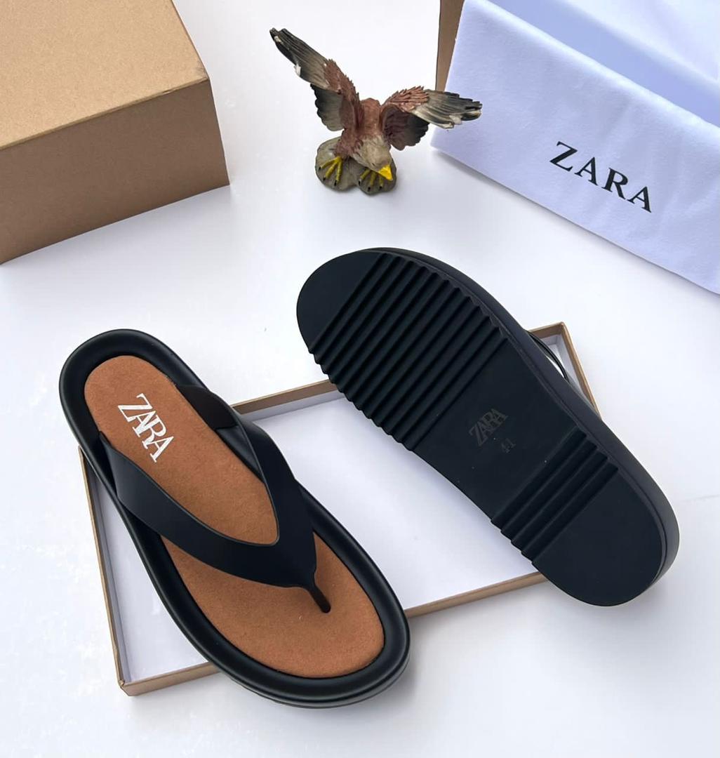 A pair of black T-strap flip flops with brown insole and rubber sole, placed on a white surface with a shoe box and bag in the background.