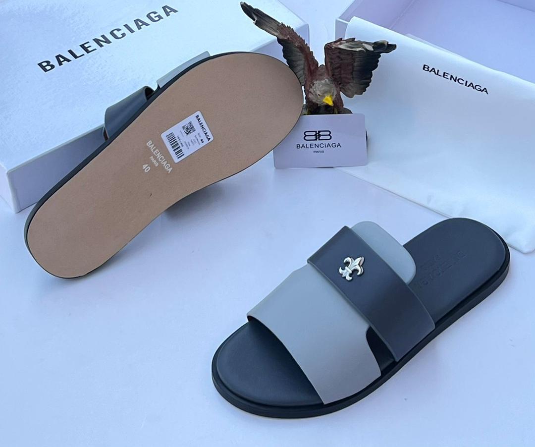 A pair of black and grey Balenciaga slide sandals with a silver logo, displayed with the brand's packaging and a decorative eagle statue.