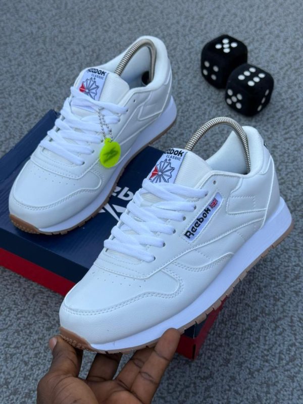 A pair of white Reebok Classic trainers with brown soles, displayed on a grey surface with a shoe box and dice in the background.