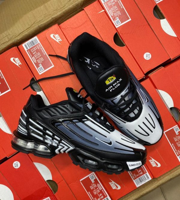 A pair of black and white Nike Air Max Plus sneakers on top of red shoe boxes, showcasing the shoe's sleek design and visible Air Max technology.