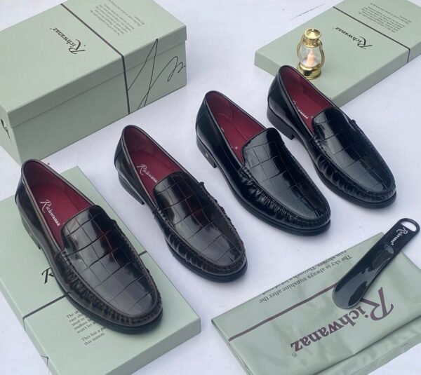 A pair of black crocodile pattern loafers with red interior lining, placed on a shoe box with a luxurious background.