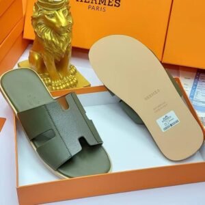 A pair of black leather Hermes Paris slide sandals in an orange box with a golden lion statue, showcasing luxury and style.