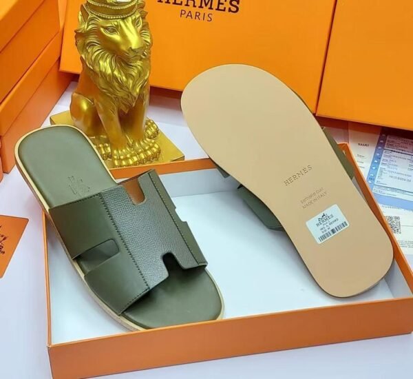 A pair of black leather Hermes Paris slide sandals in an orange box with a golden lion statue, showcasing luxury and style.