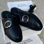 A pair of black leather mule shoes with a gold buckle, placed on top of an Alexander McQueen box and packaging, showcasing the shoe's sleek design and premium quality.
