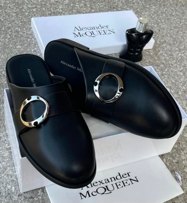 A pair of black leather mule shoes with a gold buckle, placed on top of an Alexander McQueen box and packaging, showcasing the shoe's sleek design and premium quality.