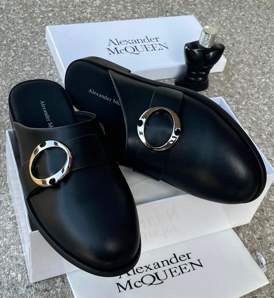 A pair of black leather mule shoes with a gold buckle, placed on top of an Alexander McQueen box and packaging, showcasing the shoe's sleek design and premium quality.