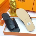A pair of black leather Hermes Paris slide sandals in an orange box with a golden lion statue, showcasing luxury and style.