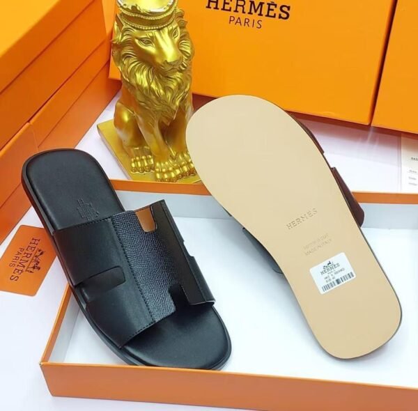 root A pair of black leather Hermes Paris slide sandals in an orange box with a golden lion statue, showcasing luxury and style.