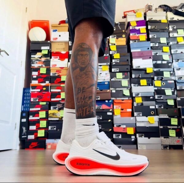 Close-up of a man's legs wearing white Nike ZoomX shoes with a black swoosh and red sole, standing in front of a collection of shoe boxes.