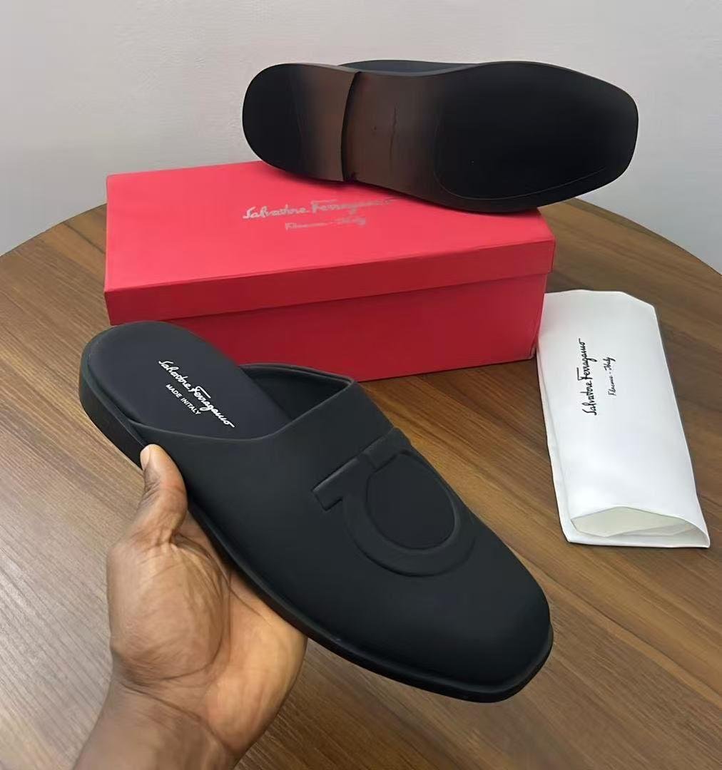 A pair of black Salvatore Ferragamo loafers on a wooden table with a red box and white dust bag, showcasing the shoe's sleek design and luxurious packaging.