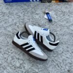 Luxury Adidas Samba sneakers in white, black, and brown, displayed on a marble surface with designer shoe boxes in the background.