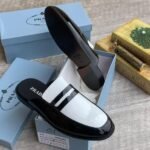 A pair of black patent leather loafers with white accent and PRADA logo, placed in a blue shoe box with a white interior.