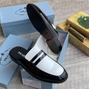 A pair of black patent leather loafers with white accent and PRADA logo, placed in a blue shoe box with a white interior.