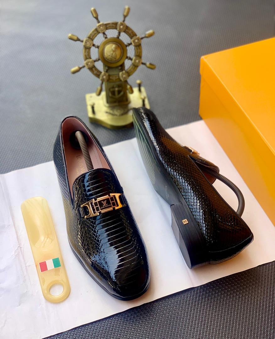 A pair of black patent leather loafers with a gold buckle on a white background, with a shoe horn and shoe box nearby.