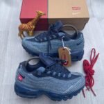 A pair of denim blue Nike Air Max sneakers with red accents and tags, accompanied by a red shoe box, a giraffe toy, and extra red laces on a light grey wooden surface.