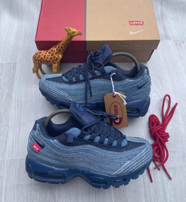 A pair of denim blue Nike Air Max sneakers with red accents and tags, accompanied by a red shoe box, a giraffe toy, and extra red laces on a light grey wooden surface.