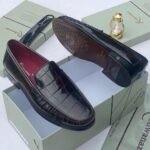 A pair of black crocodile pattern loafers with red interior lining, placed on a shoe box with a luxurious background.