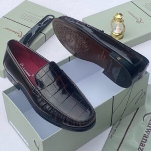 A pair of black crocodile pattern loafers with red interior lining, placed on a shoe box with a luxurious background.