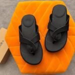A pair of black two-strap flat sandals on an orange hexagonal pillow, with a shoe box in the background, perfect for stylish women in Nigeria.