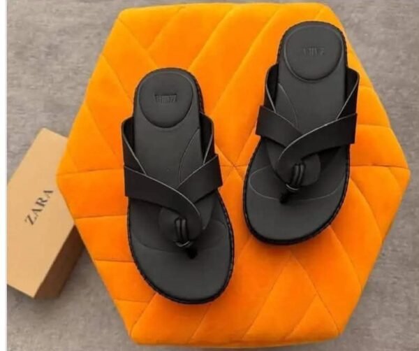 A pair of black two-strap flat sandals on an orange hexagonal pillow, with a shoe box in the background, perfect for stylish women in Nigeria.