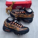 A pair of camouflage Nike Air Max sneakers with orange accents, displayed with a giraffe toy and a red Nike box on a light grey wooden floor.