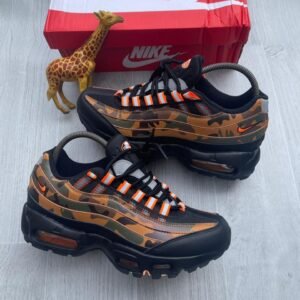 A pair of camouflage Nike Air Max sneakers with orange accents, displayed with a giraffe toy and a red Nike box on a light grey wooden floor.