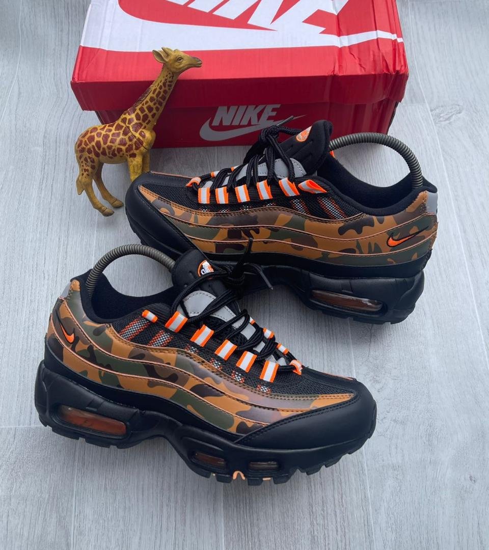 A pair of camouflage Nike Air Max sneakers with orange accents, displayed with a giraffe toy and a red Nike box on a light grey wooden floor.