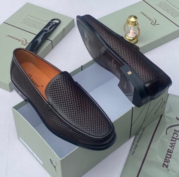 A pair of black woven leather loafers displayed on an open shoe box with accessories, showcasing the shoe's intricate design and luxurious texture.