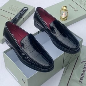 A pair of black crocodile pattern loafers with red interior lining, placed on a shoe box with a luxurious background.