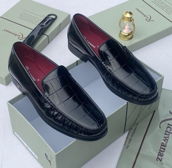 A pair of black crocodile pattern loafers with red interior lining, placed on a shoe box with a luxurious background.