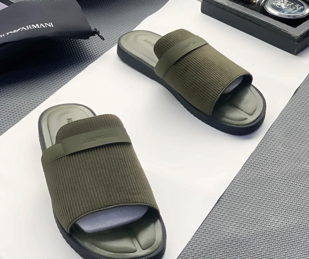 A pair of green ribbed slide sandals from Armani, displayed on a white surface with a black pouch and watch in the background.