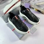 A pair of black Prada sneakers with red accents and white soles, placed on top of a Prada shoe box with luxury fashion books in the background.