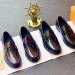 A stylish display of four black leather loafers on a white paper, accompanied by a gold ship's wheel and an orange shoe box, showcasing the product's sleek design and premium quality.