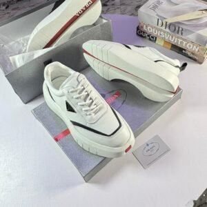 A pair of white Prada sneakers with red accents, placed on top of their box with a luxury brand book in the background.