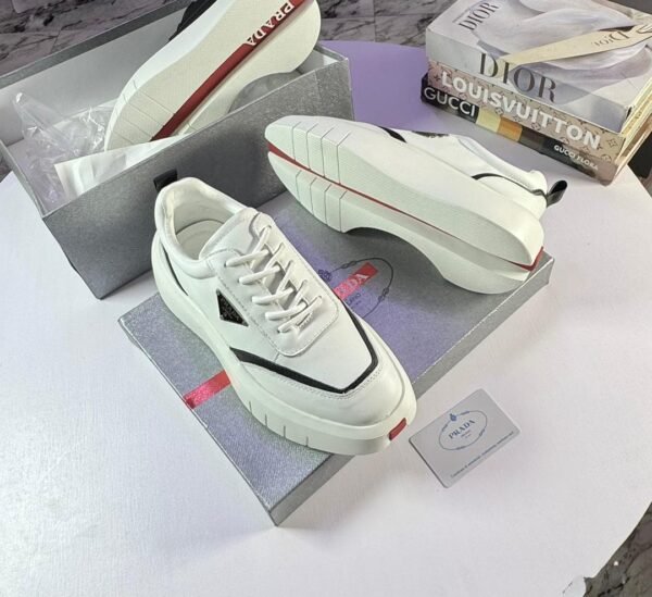 A pair of white Prada sneakers with red accents, placed on top of their box with a luxury brand book in the background.