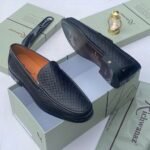 A pair of black woven leather loafers displayed on an open shoe box with accessories, showcasing the shoe's intricate design and luxurious texture.