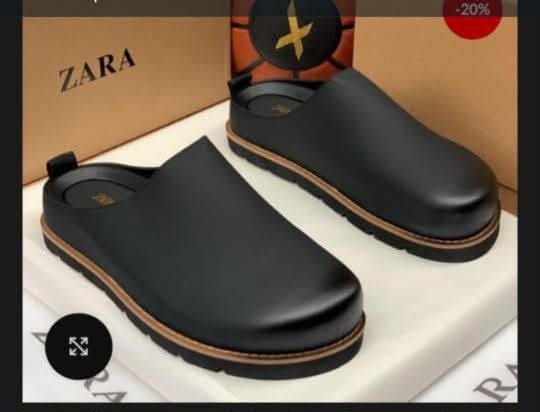 Close-up of black leather clog shoes on a white box with Zara branding in the background, showcasing stylish and comfortable footwear for men.