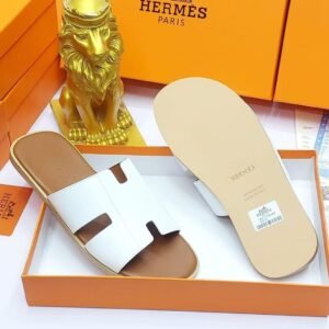 A pair of black leather Hermes Paris slide sandals in an orange box with a golden lion statue, showcasing luxury and style.