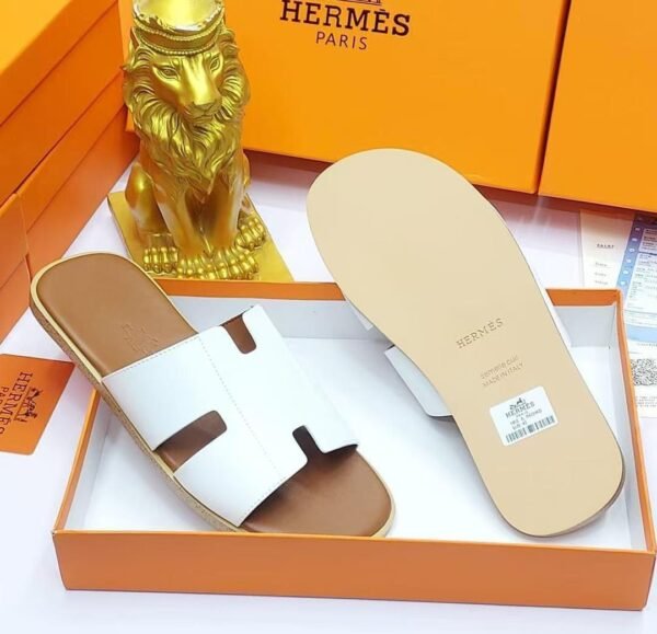A pair of black leather Hermes Paris slide sandals in an orange box with a golden lion statue, showcasing luxury and style.
