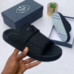 A pair of black Prada slides with a comfortable design and stylish logo, displayed with a box and a hand holding the slide.