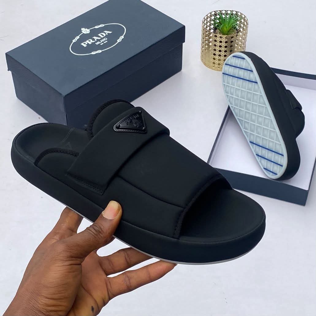 A pair of black Prada slides with a comfortable design and stylish logo, displayed with a box and a hand holding the slide.