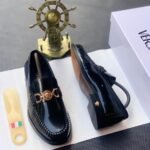 A pair of black leather loafers with gold buckle, placed on a white shoe box with a shoe horn and a decorative ship's wheel in the background.