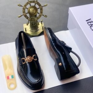 A pair of black leather loafers with gold buckle, placed on a white shoe box with a shoe horn and a decorative ship's wheel in the background.