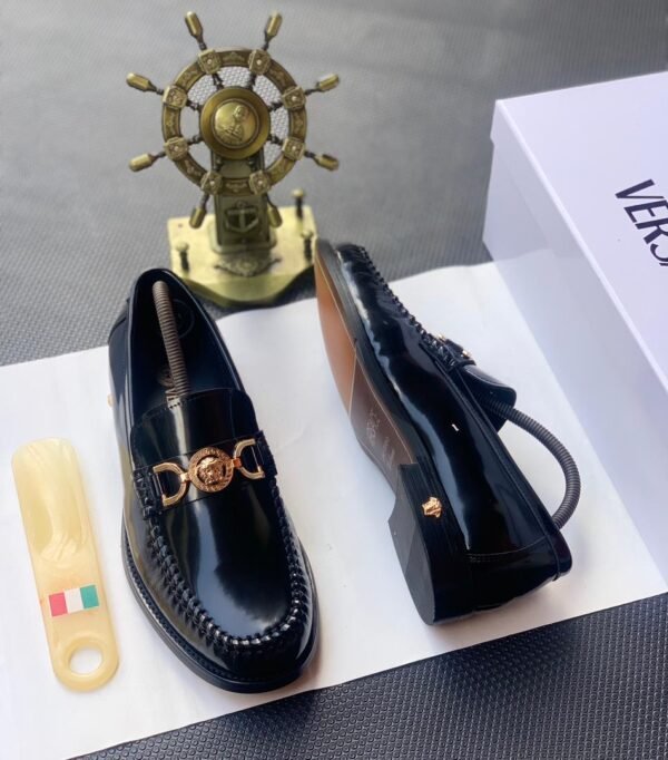 A pair of black leather loafers with gold buckle, placed on a white shoe box with a shoe horn and a decorative ship's wheel in the background.