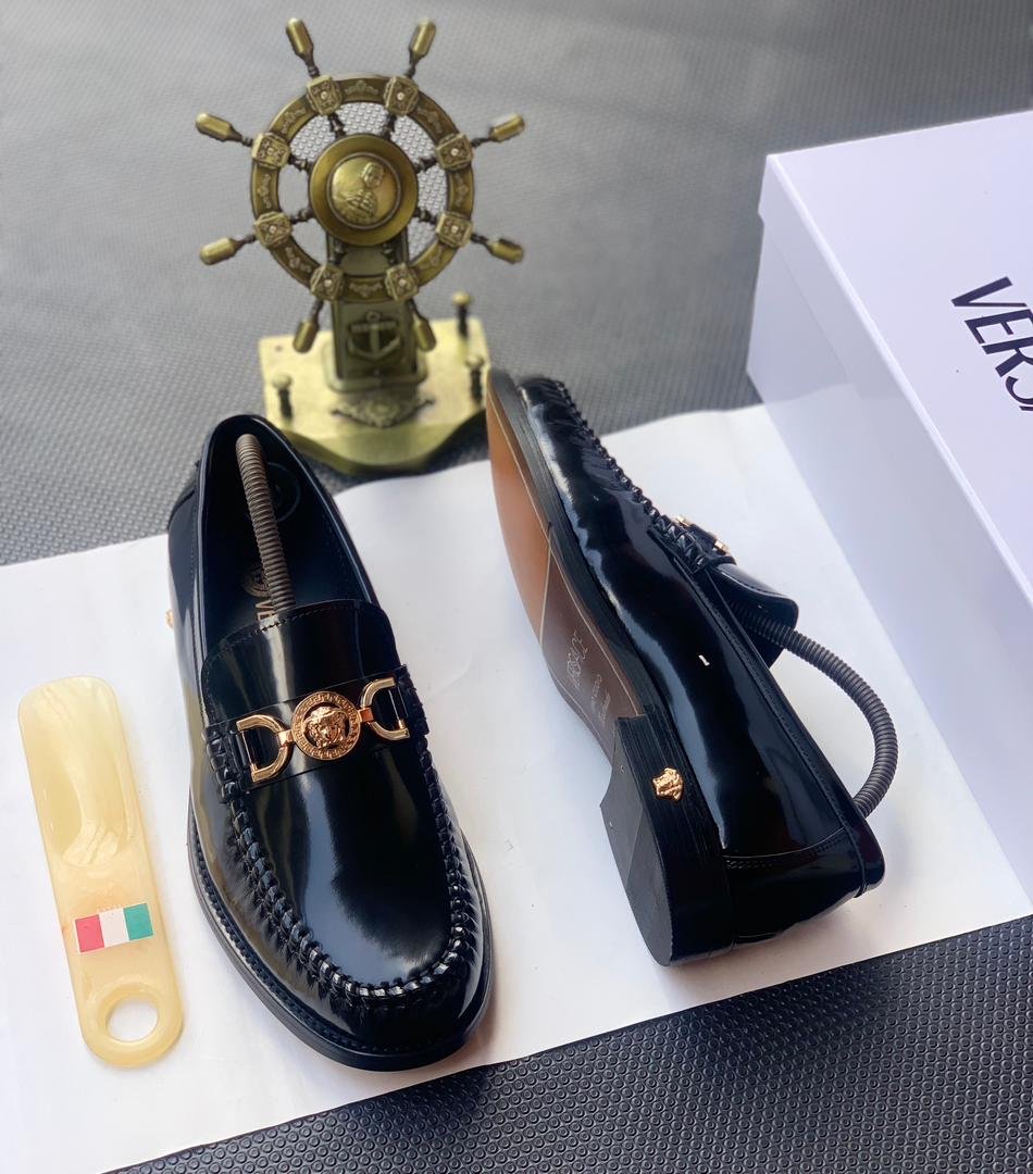 A pair of black leather loafers with gold buckle, placed on a white shoe box with a shoe horn and a decorative ship's wheel in the background.
