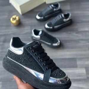 A close-up of a hand holding a black rhinestone sneaker with silver accents, with two more pairs in the background on a grey wooden floor.